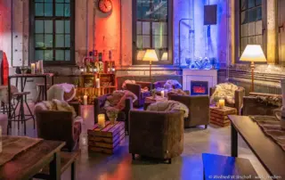 Sitzecken aus braunen Ledersesseln mit Fellen und Kaminfeuer in einer rustikalen Industriehalle.